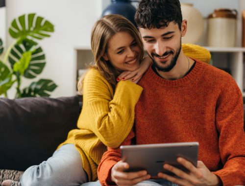 Pareja heterosexual sentada en el sofá de su casa viendo algo en su tablet.