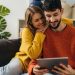 Pareja heterosexual sentada en el sofá de su casa viendo algo en su tablet.