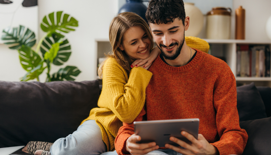 Pareja heterosexual sentada en el sofá de su casa viendo algo en su tablet.