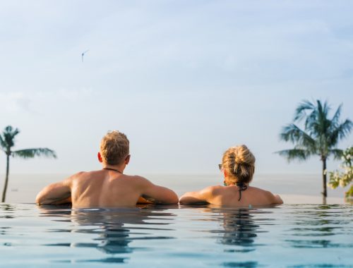Pareja heterosexual dentro de una alberca de borde infinito frente a una hermosa vista con palmeras grandes.
