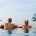 Pareja heterosexual dentro de una alberca de borde infinito frente a una hermosa vista con palmeras grandes.