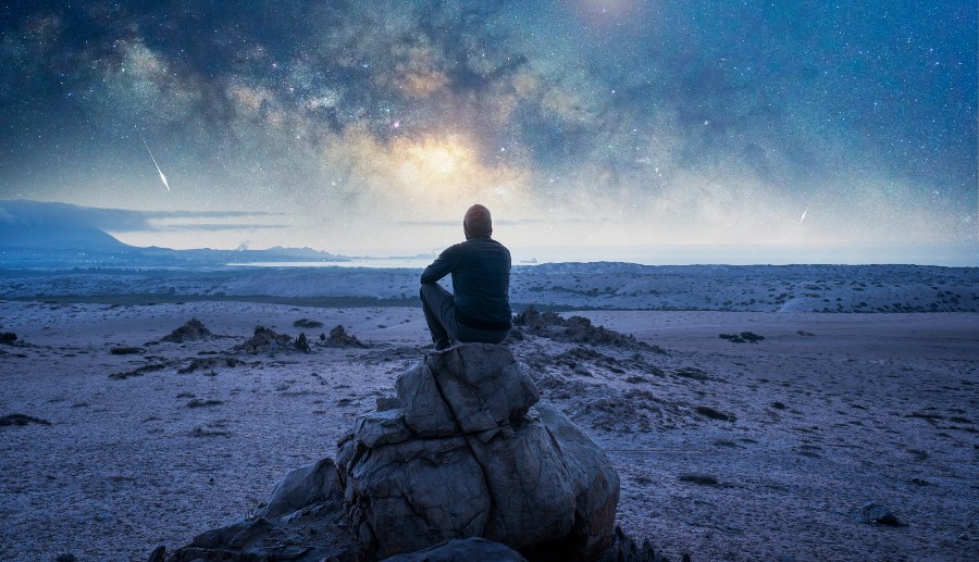 Hombre sentado sobre una roca observando el cielo colorido con lluvia de meteoros