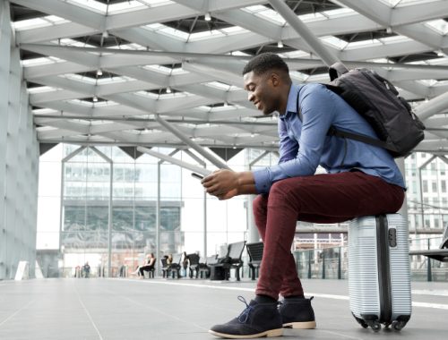 Hombre negro sentado sobre su propia maleta en un aeropuerto usando su celular