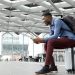 Hombre negro sentado sobre su propia maleta en un aeropuerto usando su celular