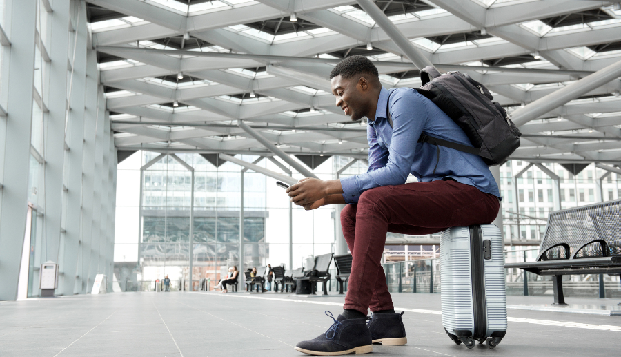 Hombre negro sentado sobre su propia maleta en un aeropuerto usando su celular